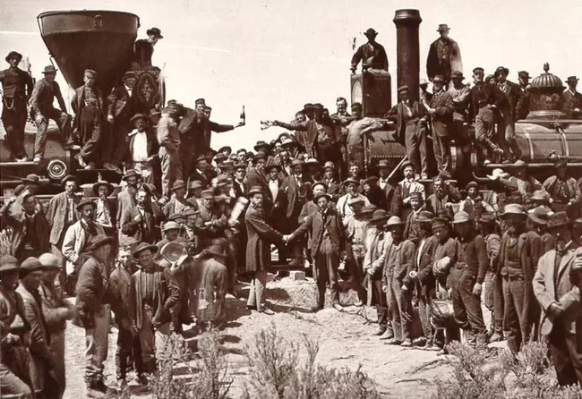 A celebratory photo taken at the point the eastern and western sections of the transcontinental railroad met. The photo shows two train engines facing each other. One man stands at the front of each engine--one holding a bottle of champagne and one holding a champagne glass. Dozens of others stand in front of or on top of the train engines.