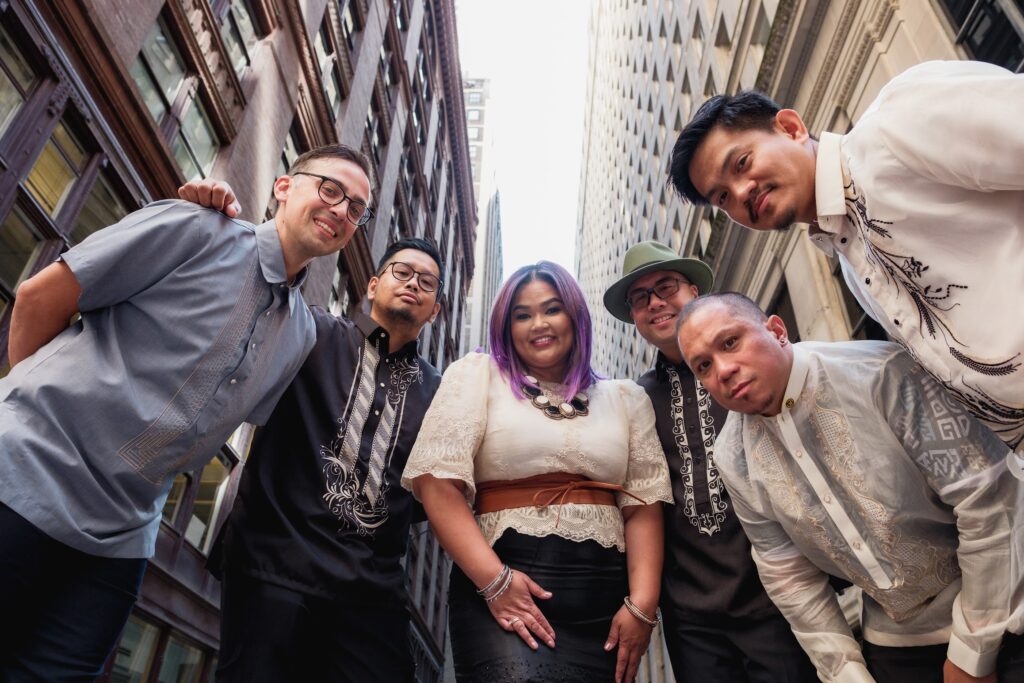 Publicity Photo of The Sama Sama Project. The members are standing in a semi-circle with hi-rises in the background.